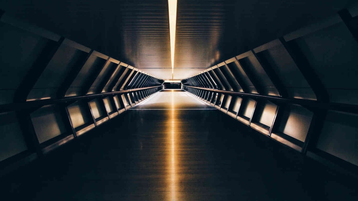 A futuristic, tunnel-like corridor with a glowing strip of light on the ceiling, leading to a bright exit in the distance. The walls and floor are dark with reflective surfaces.