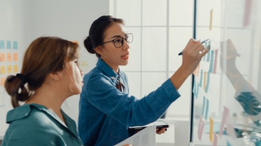 Two Young Asian businesswomen in smart casual wear writing on colorful sticky notes on a whiteboard